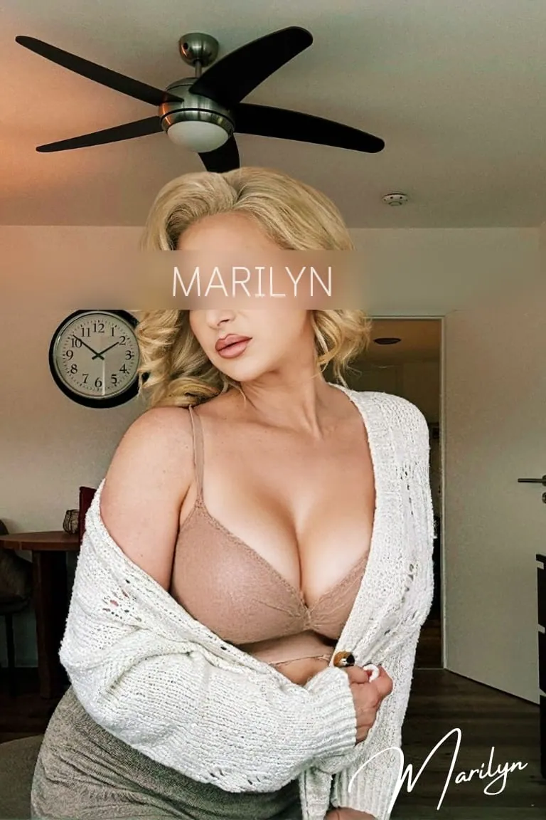 Marilyn in beigem BH und cremefarbener Strickjacke vor einer Wanduhr