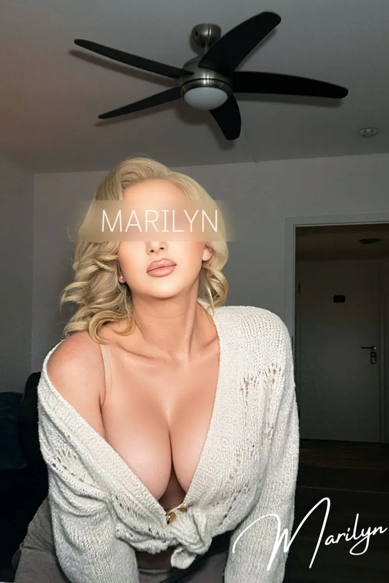 Marilyn in offener cremefarbener Strickjacke, nach vorn geneigt unter einem Deckenventilator