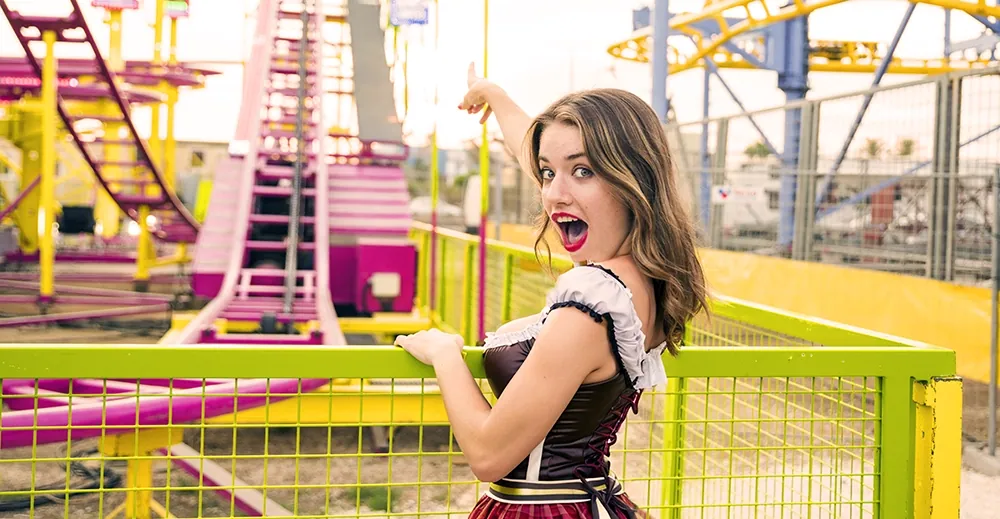 Eine junge Frau im Dirndl zeigt auf dem Cannstatter Volksfest begeistert auf eine Achterbahn