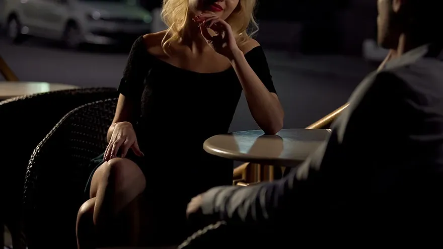 Eine blonde Frau im schwarzen Kleid sitzt abends einem Mann an einem kleinen Bistrotisch gegenüber