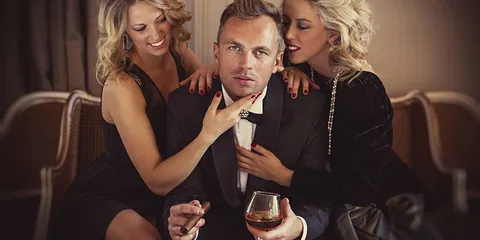 Ein Mann im Smoking mit Zigarre und Cognacglas zwischen zwei blonden Frauen auf einem Sofa
