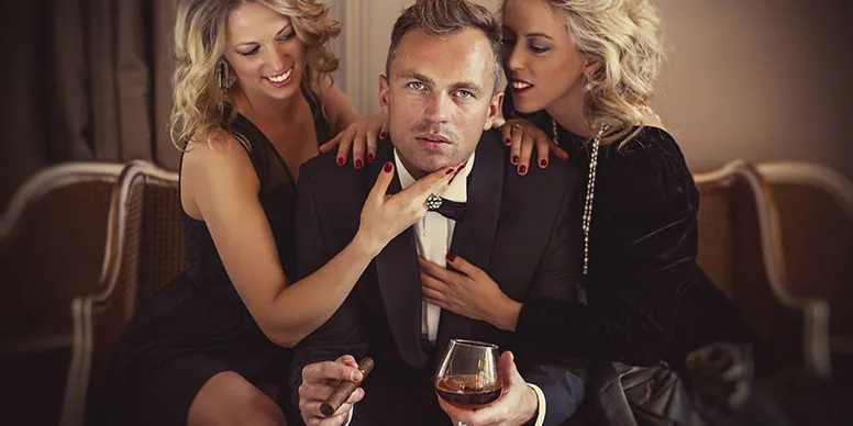 Ein Mann im Smoking mit Zigarre und Cognacglas zwischen zwei blonden Frauen auf einem Sofa
